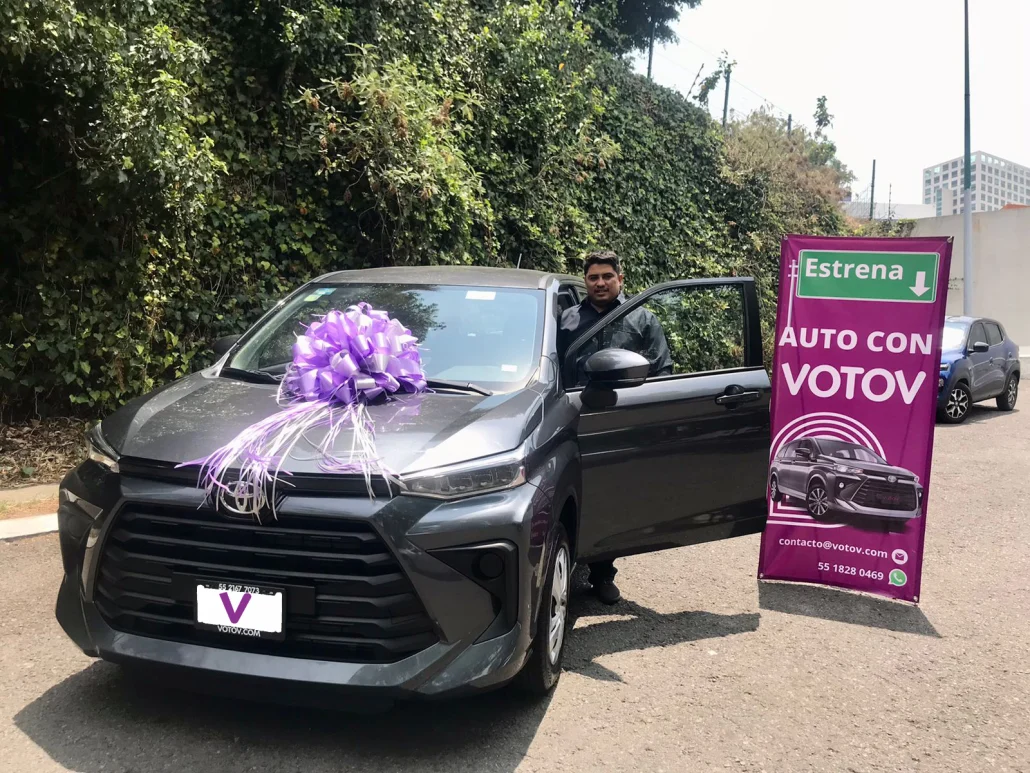 VOTOVer en la puerta de su Toyota Avanza gris nueva con moño lila, junto al banner de VOTOV