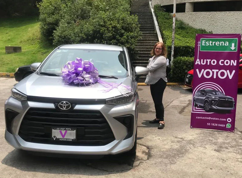 VOTOVer junto a su Toyota Avanza plata nueva con moño lila en el día de su entrega