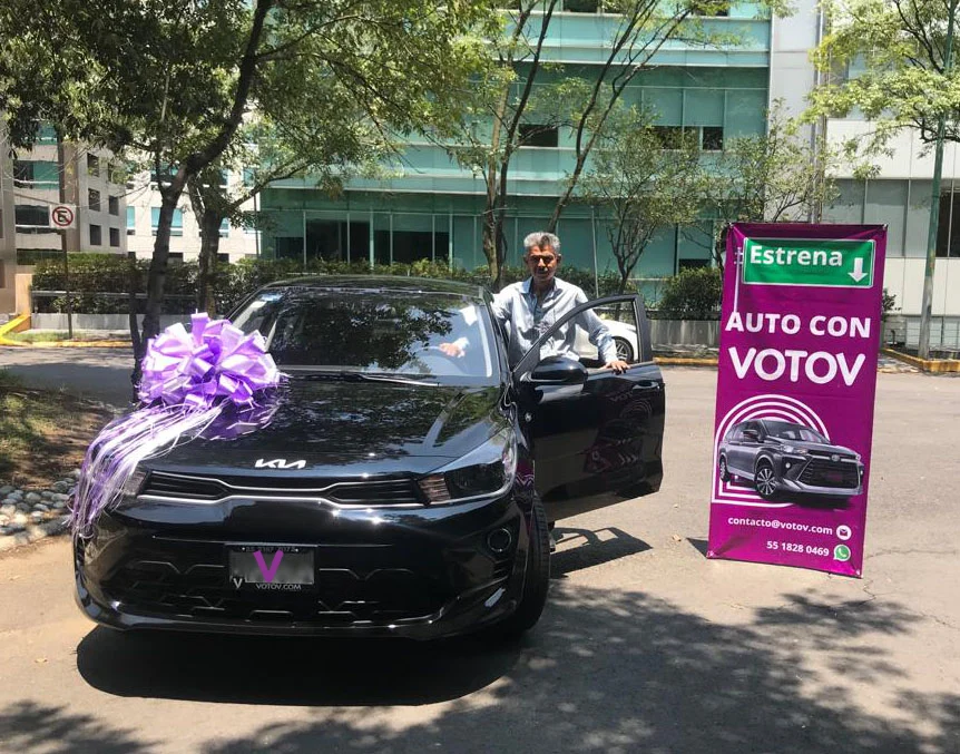 VOTOVer junto a su Kia sedán negro nuevo con moño lila, al lado del banner Estrena auto con VOTOV