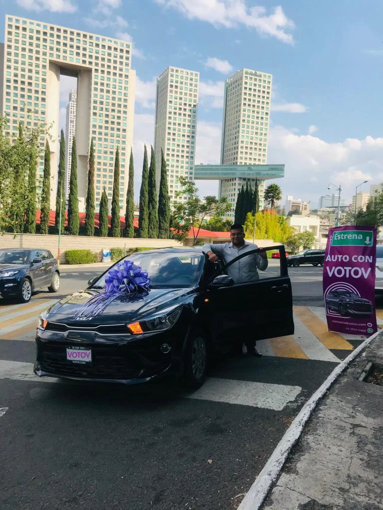 VOTOVer recargado en la puerta abierta de su Kia sedán negro con moño azul, frente al edificio de los Arcos Bosques en la Ciudad de México